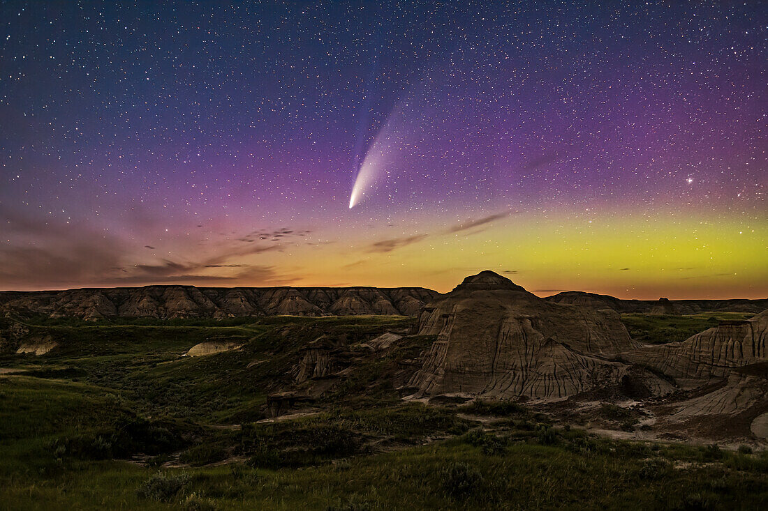 Dies ist der Komet NEOWISE (C/2020 F3) über den Badlands und Formationen des Dinosaur Provincial Park, Alberta, in der Nacht vom 14. auf den 15. Juli 2020 um etwa 12:30 Uhr Ortszeit, als er fast genau nördlich und so tief wie möglich in dieser Nacht auf 51° nördlicher Breite stand. Ein grünes und magentafarbenes Polarlicht färbt den nördlichen Himmel ebenfalls blau mit ewiger Sommerdämmerung. Capella ist ganz rechts zu sehen.