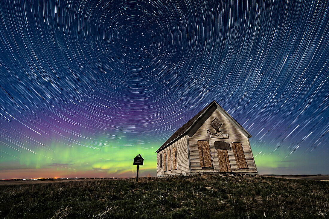 Das Liberty Schoolhouse von 1910, eine klassische Einraumschule der Pioniere, in der Prärie von Alberta unter dem Sternenhimmel einer Frühlingsnacht, mit zirkumpolaren Sternenspuren, die den Polarstern umkreisen, und einer tanzenden Aurora im Norden. Das Mondlicht des 8 Tage alten zunehmenden Mondes sorgt für die Beleuchtung.