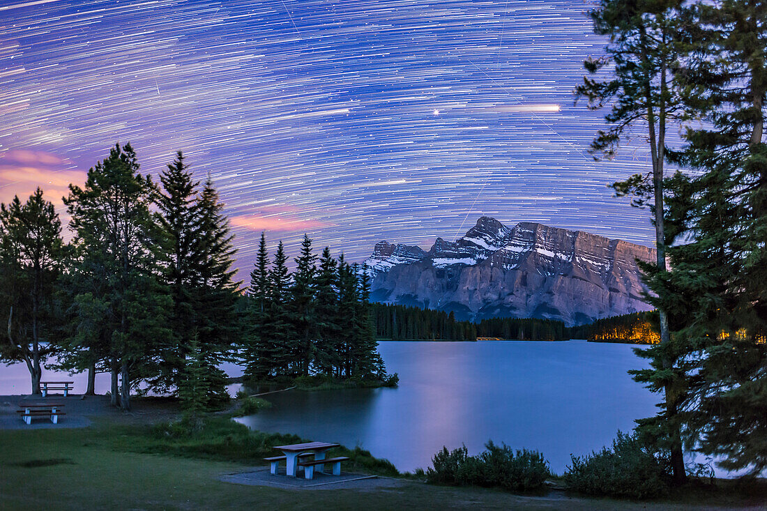 Die Sterne und der Mars im Schlepptau über Mt. Rundle und Two Jack Lake im Banff National Park, Alberta, am 3. Juni 2016. Mars ist der hellste Streifen und das hellste Objekt am Himmel. Beachten Sie den Glitzerpfad des Mars im Wasser. Satelliten streifen durch die Sternenspuren.
