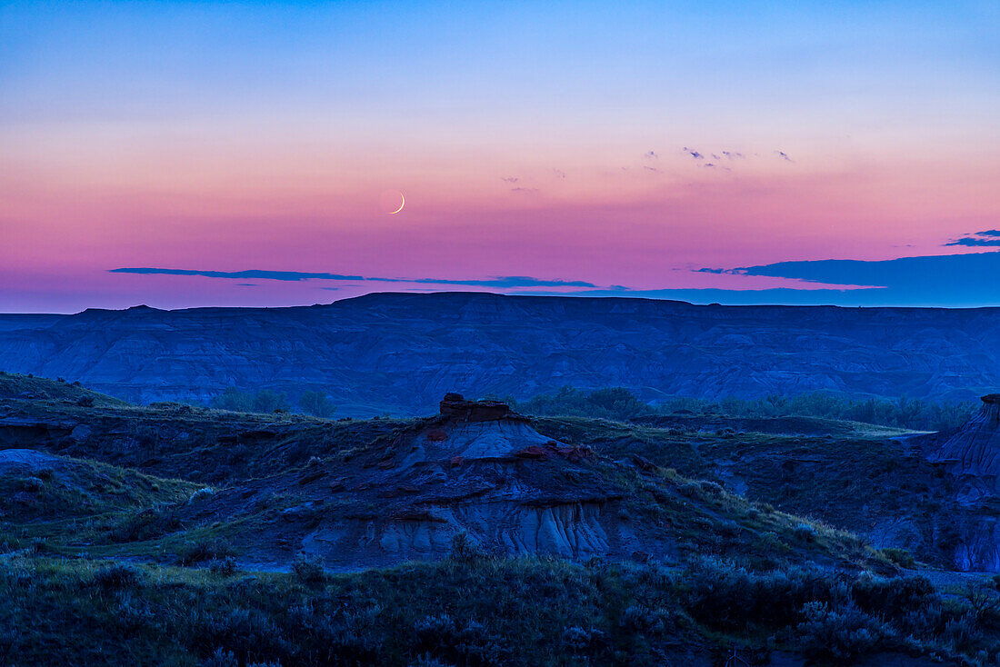 Die zunehmende Mondsichel zwei Tage nach Neumond an einem rauchigen Himmel über den Badlands des Dinosaur Provincial Park, Alberta, Kanada. Dies war am 30. Juni 2022. Trotz des dunstigen Himmels sind die Farben der Dämmerung am Himmel gut zu erkennen. Auf der Nachtseite des Mondes ist das Glühen des Erdscheins leicht zu erkennen.