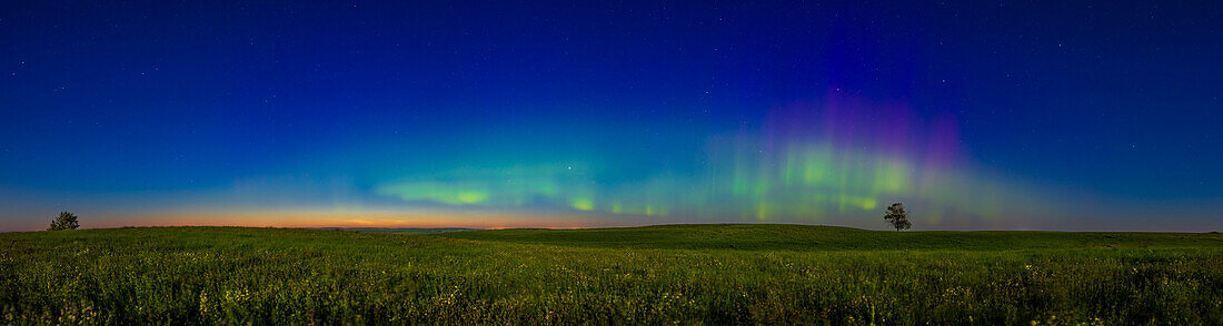An arc of modest aurora borealis (Northern Lights) across the northern horizon, above a prairie meadow in the light of a bright waxing gibbous Moon, a day before Full. This was just after midnight on July 11/12, 2022 when the sky was also still lit by summer twilight colours, and with a weak display of noctilucent clouds also visible low in the northwest at left. The auroral curtains exhibit the usual green band and rays, but also upper altitude reds and purples and a faint blue tint at the very tops where the aurora is lit by the Sun. The aurora was never bright this night (Kp Index was 3 or 