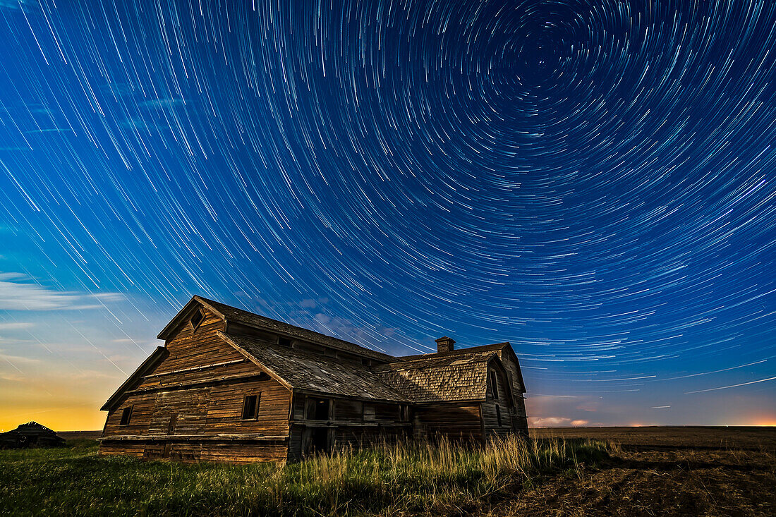 Zirkumpolarer Sternenschweif über einer großen alten Scheune in Süd-Alberta, in einer schönen Frühlingsnacht, 23. Mai 2018. Die Beleuchtung erfolgt durch den zunehmenden Gibbous-Mond im Süden. Der Blick geht nach Norden zum Polarstern oben rechts. Am nördlichen Horizont ist ein Gewitter mit einem Blitz als Bonus zu sehen.