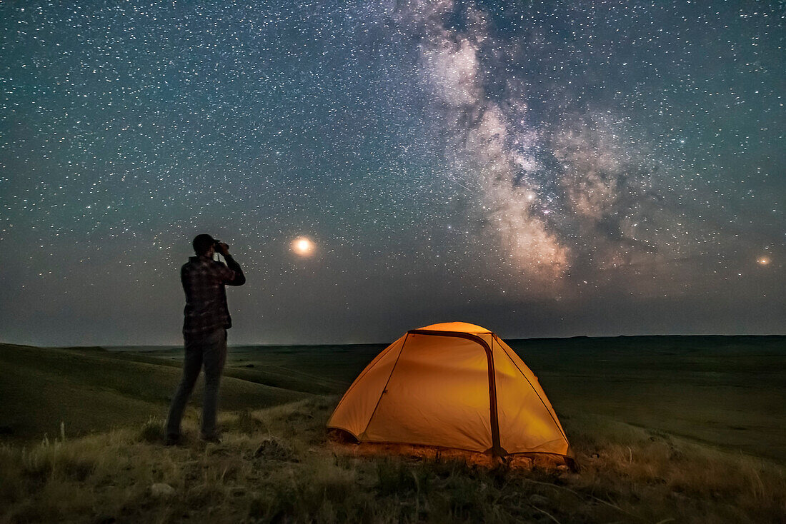 Ein Parkdolmetscher posiert für eine Szene im Grasslands National Park, Saskatchewan, bei der Sternenbeobachtung mit einem Fernglas unter der Milchstraße in einer dunklen mondlosen Nacht. Grasslands eignet sich perfekt für die Sternenbeobachtung, da es ein Dark Sky Preserve ist und der Horizont weit und unverbaut ist.