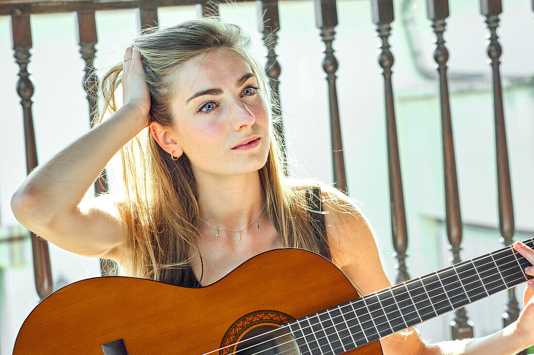 Porträt einer jungen schönen kaukasischen Frau in den 20ern mit langen Haaren und blauen Augen, die auf der Veranda eines Landhauses Gitarre spielt. Lebensstil-Konzept.