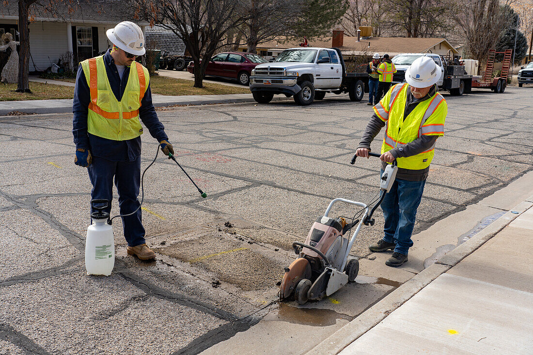 Arbeiter eines Versorgungsunternehmens entfernen mit einer Säge einen Teil des Straßenbelags, um eine Versorgungsleitung unter der Straße zu reparieren. Ein zweiter Arbeiter versprüht Wasser als Schmier- und Kühlmittel.