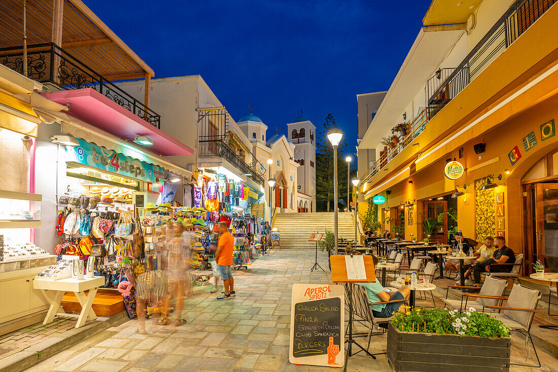 Blick auf Geschäfte und Restaurants mit der Kirche von Agia Paraskevi im Hintergrund in Kos Stadt in der Abenddämmerung, Kos, Dodekanes, Griechische Inseln, Griechenland, Europa
