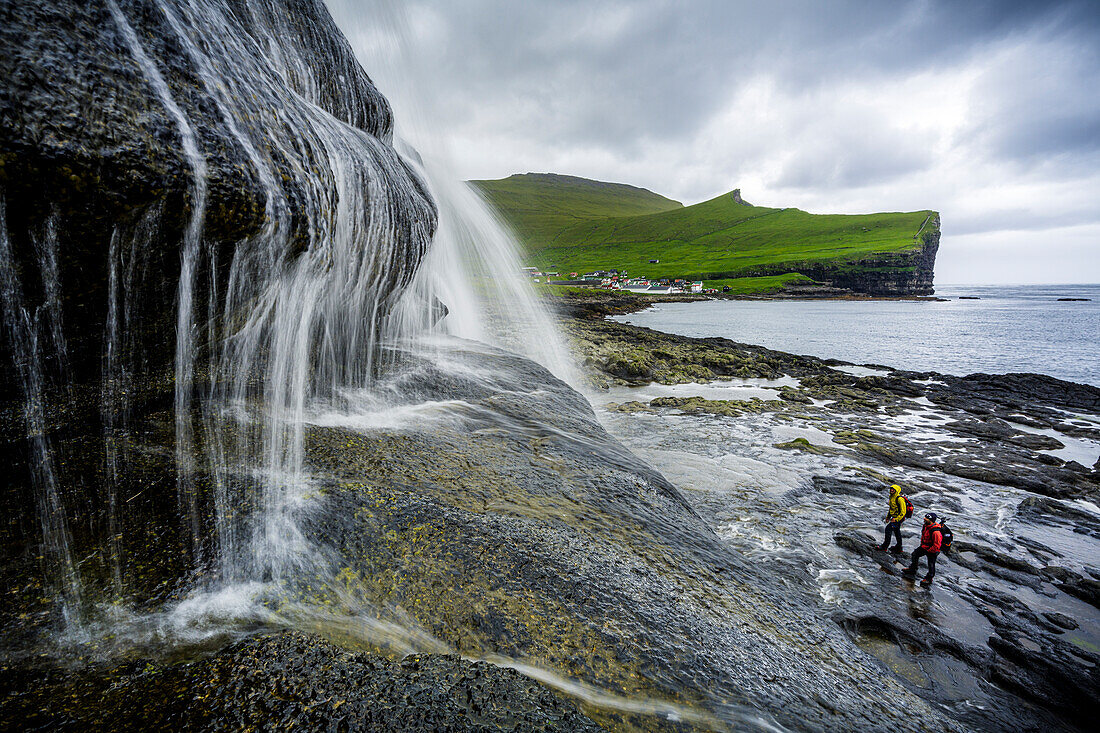 High angle view of two hikers admiring a majestic waterfall standing on cliffs, Gjogv, Eysturoy Island, Faroe Islands, Denmark, Europe