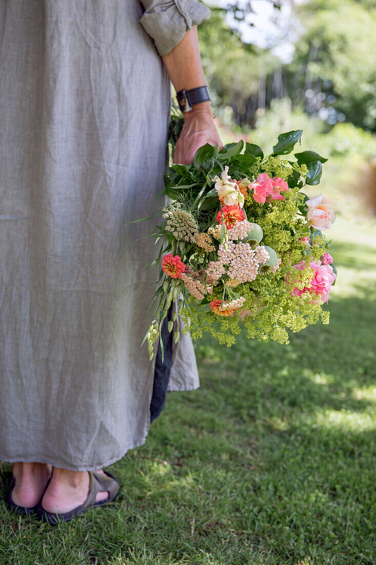 Frau in Leinenkleid hält sommerlichen Blumenstrauß im Garten