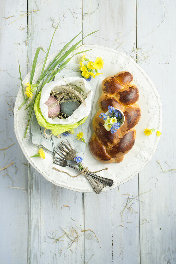 Osterzopf mit eingebackener Eierschale gefüllt mit Schlüsselblumen und Vergissmeinnicht, Ostereier in einer Ostertüte und Besteck