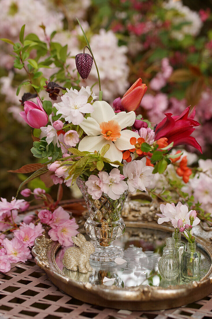 Frühlingsstrauß mit Narzisse (Narcissus), Schachbrettblume und Tulpe auf silbernem Tablett mit Mandelblüten in Parfümflacons, Zierkirschenzweig und Osterhasenfigur