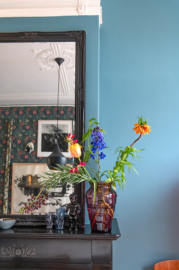 Colorful flower arrangement in a red glass vase on a black mantelpiece against a blue-painted wall