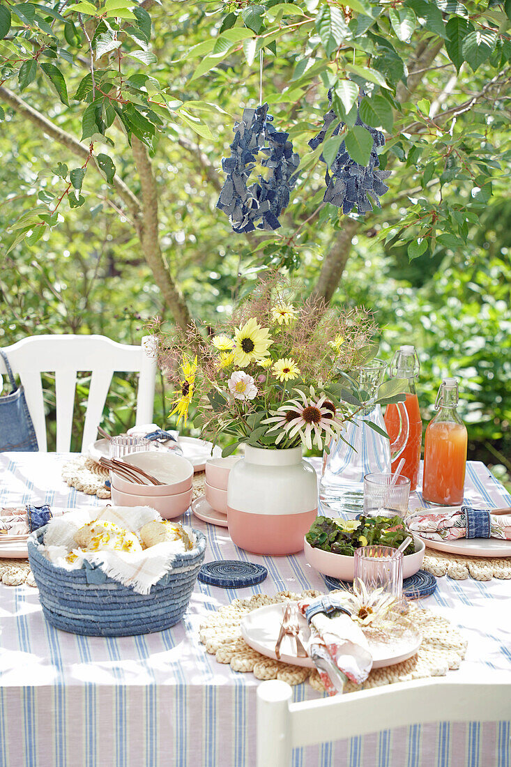 Gedeckter Gartentisch mit Blumenstrauß und Saftflaschen unter einem Baum