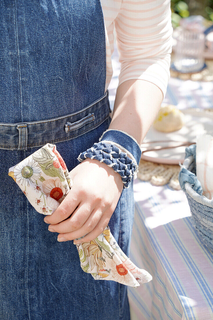 Person in Jeansschürze und Armbändern aus Jeans hält Serviette mit Blumenmuster in der Hand