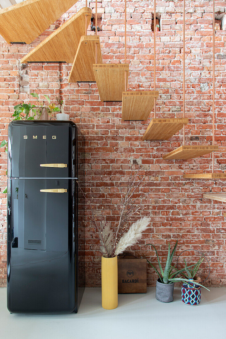 Schwarzer Retro-Kühlschrank vor freiliegender Backsteinwand mit hängender Holztreppe