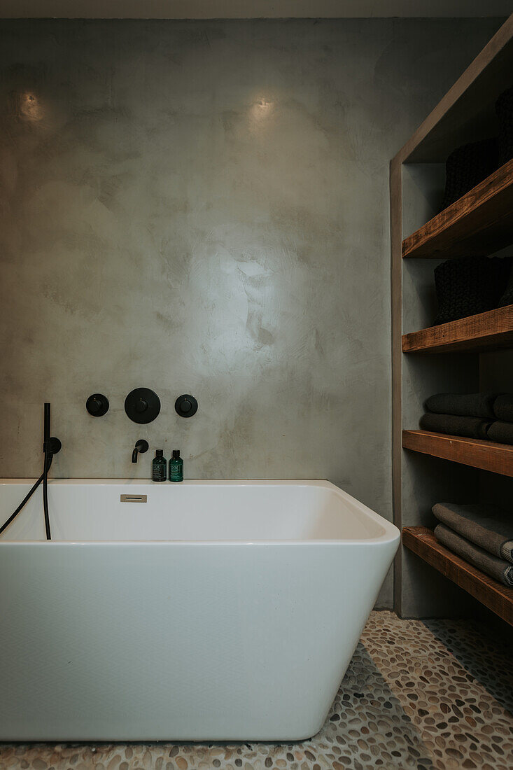 Free-standing bathtub in front of concrete wall and shelves with towels