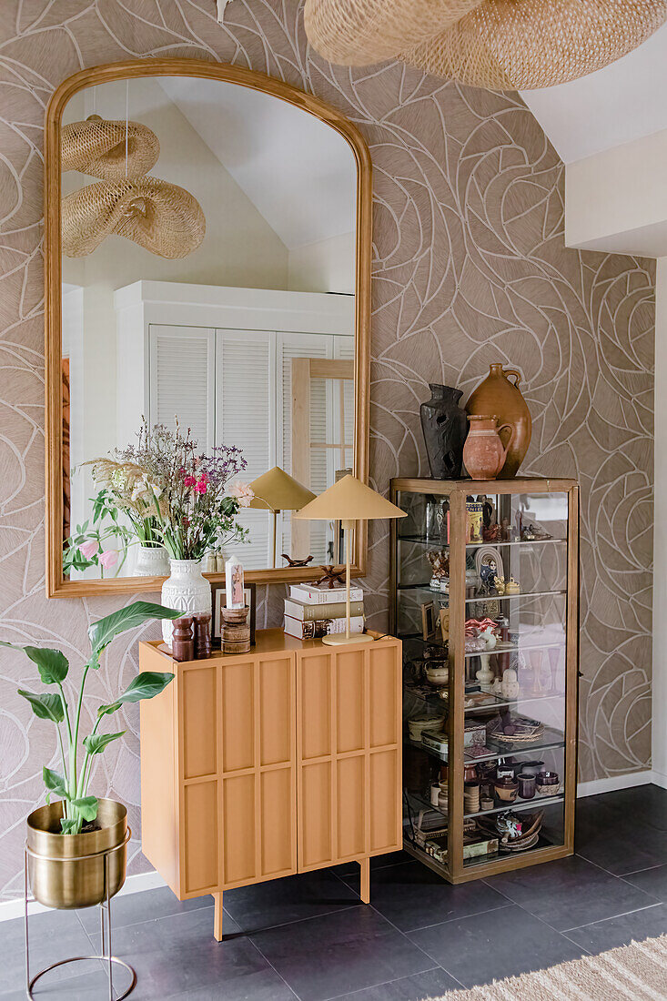 Sideboard und Glasvitrine mit Dekogegenständen im Flur vor gemusterter Tapete und großem Spiegel