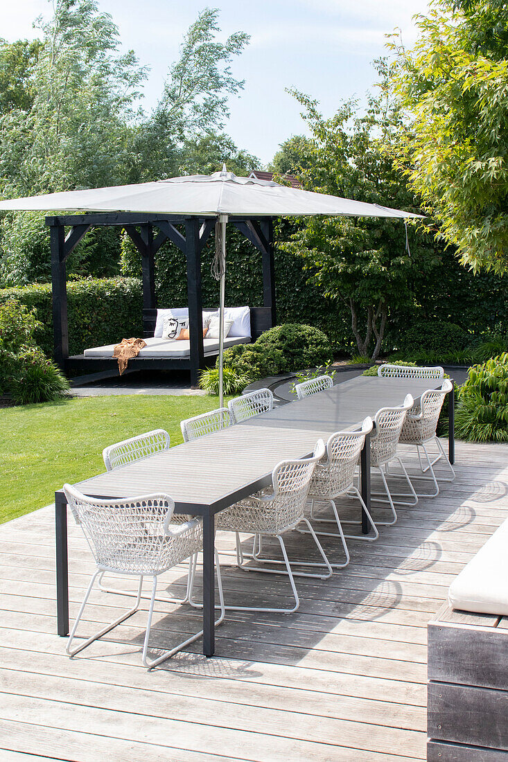 Große Holzterrasse mit Esstisch, weißen Stühlen, Sonnenschirm und Gartenpavillon im Hintergrund