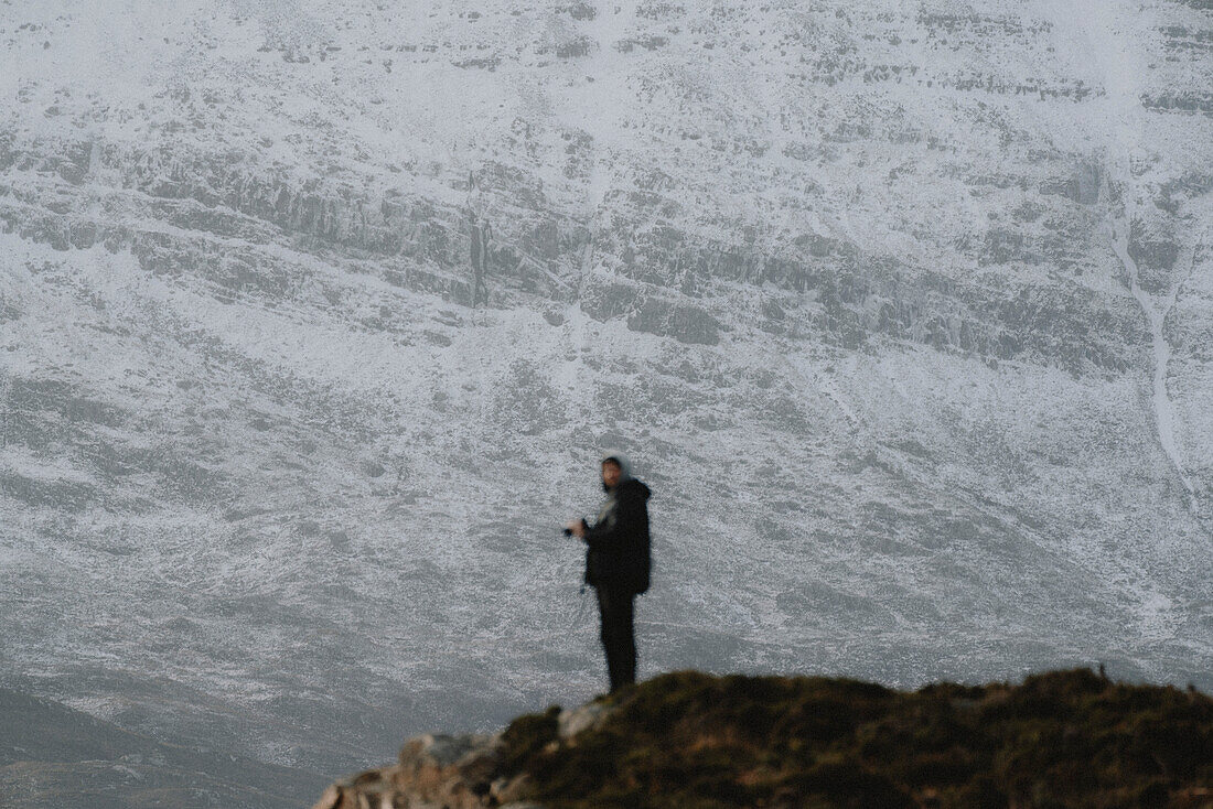 Photographer standing on hill below snowcapped mountain, Assynt, Sutherland, Scotland\n