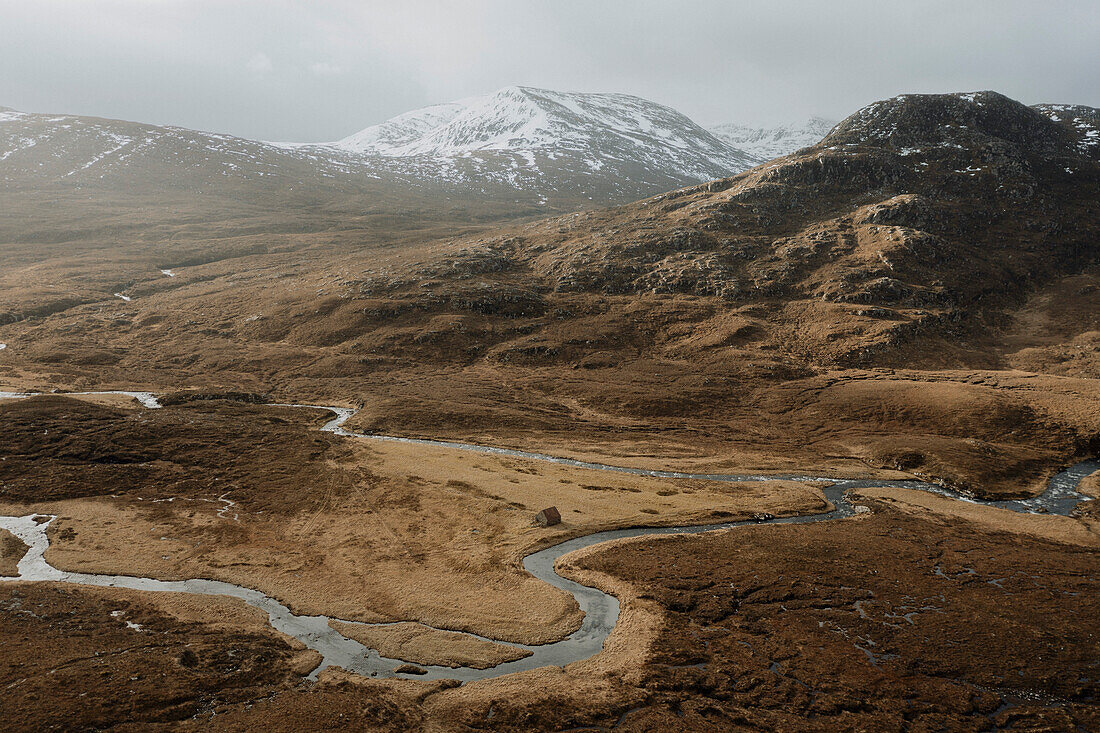 Gewundener Fluss in der Landschaft unterhalb eines schneebedeckten Berges, Assynt, Sutherland, Schottland