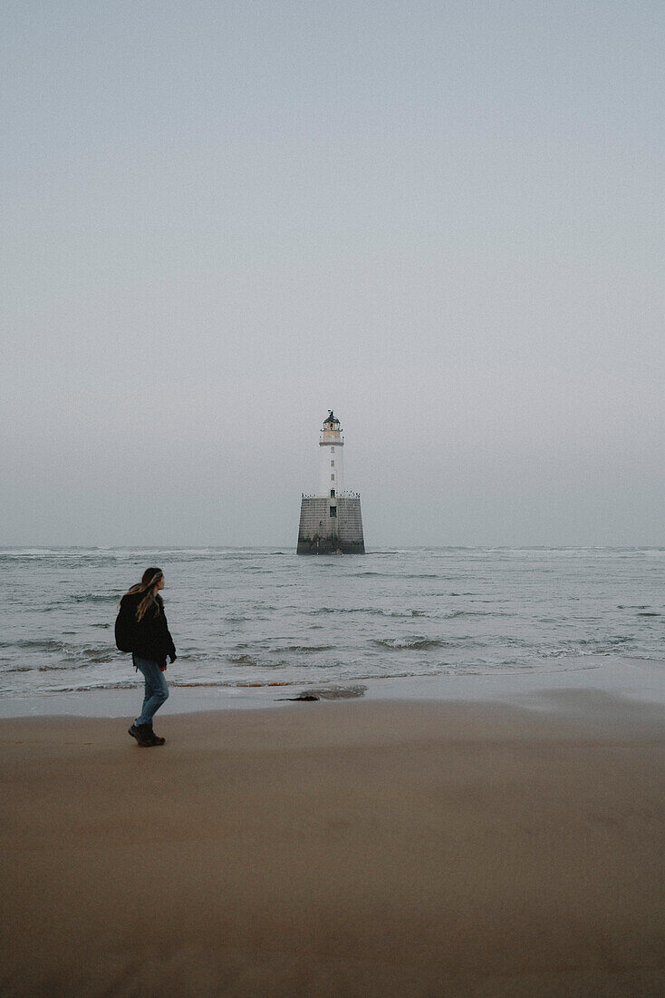 Frau spaziert am Meeresstrand mit Leuchtturm, Rattray, Aberdeenshire, Schottland