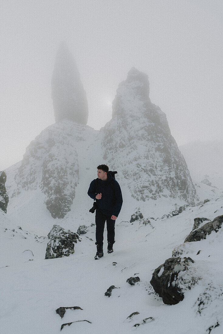 Männlicher Wanderer mit Kamera beim Wandern unterhalb einer schneebedeckten Felsformation, Old Man of Storr, Schottland
