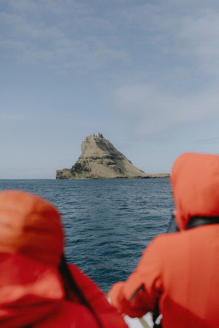 Tourists on ferry looking at rock formation over sunny ocean, Mykines, Faroe Islands\n