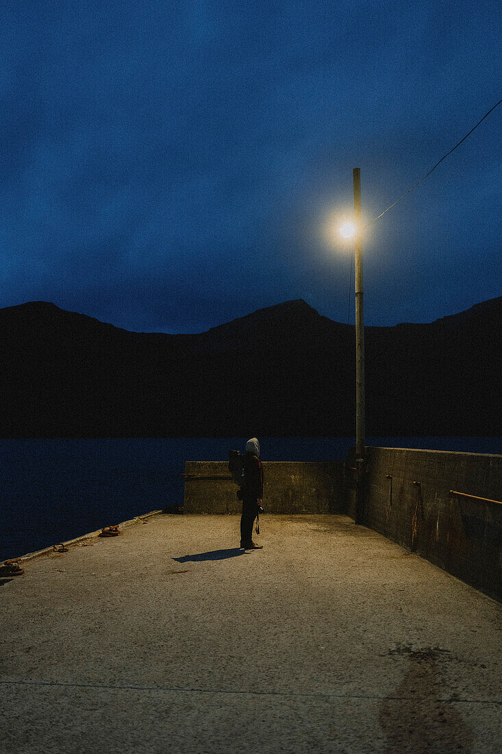 Man on dark jetty looking up at streetlamp, Kunoy, Faroe Islands\n