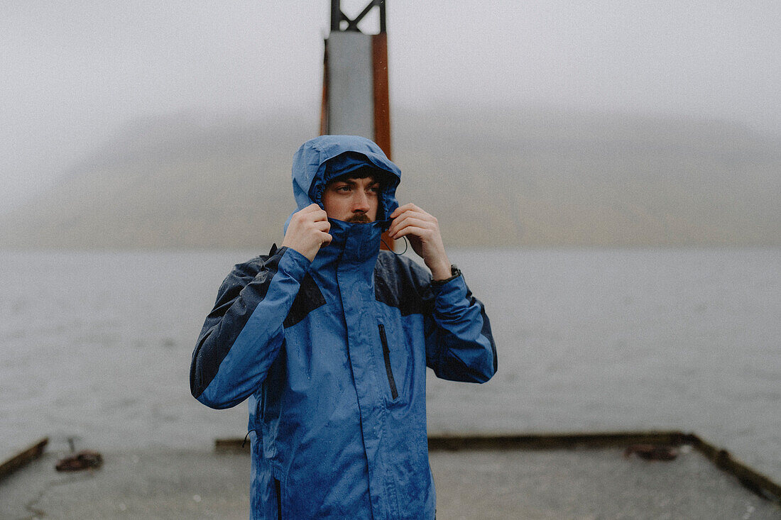 Man in hooded jacket standing on rainy jetty at ocean, Kollafjorour, Faroe Islands\n