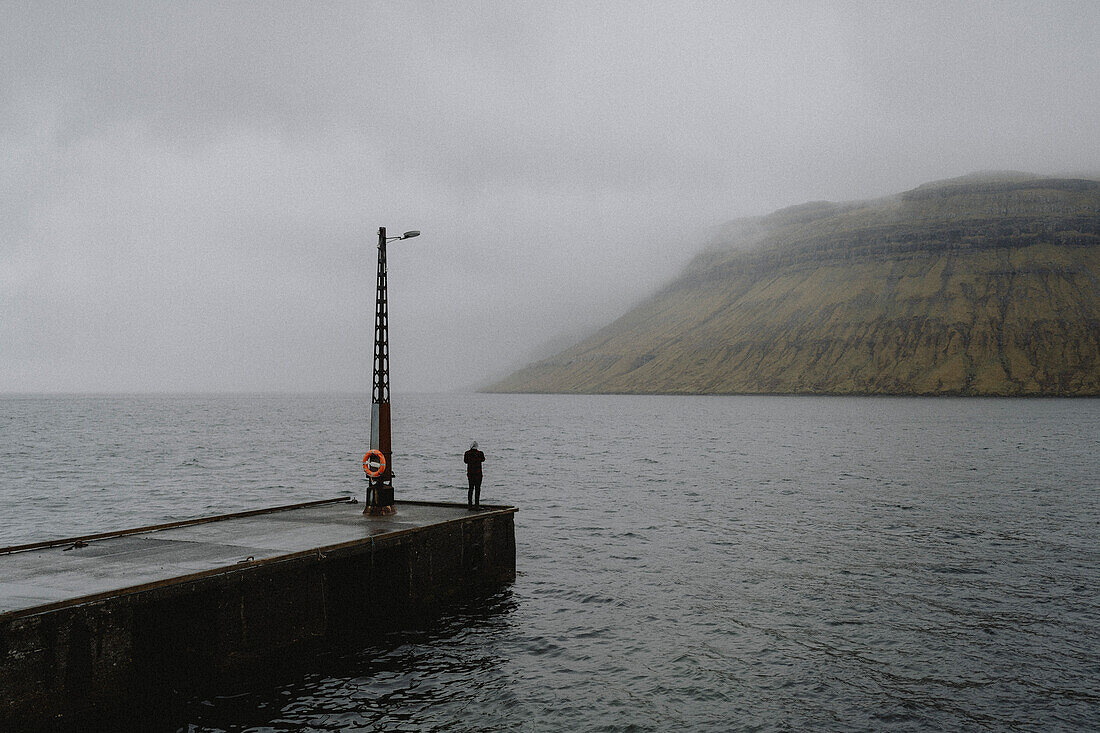Man on jetty looking at foggy ocean seascape, Kollafjorour, Faroe Islands\n