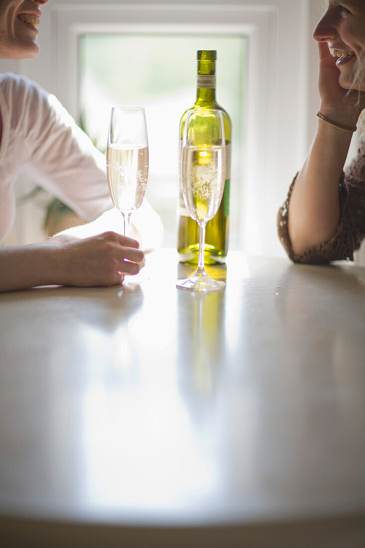 Zwei junge Frauen sitzen am Tisch, unterhalten sich lachend und trinken Wein