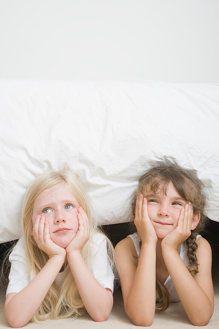 Two young girls with chin resting on hands under bed\n