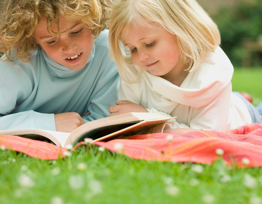 Two children lying in the garden reading a book\n