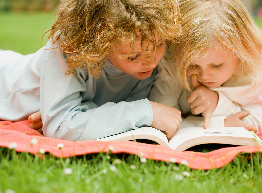 Two children lying in the garden reading a book\n
