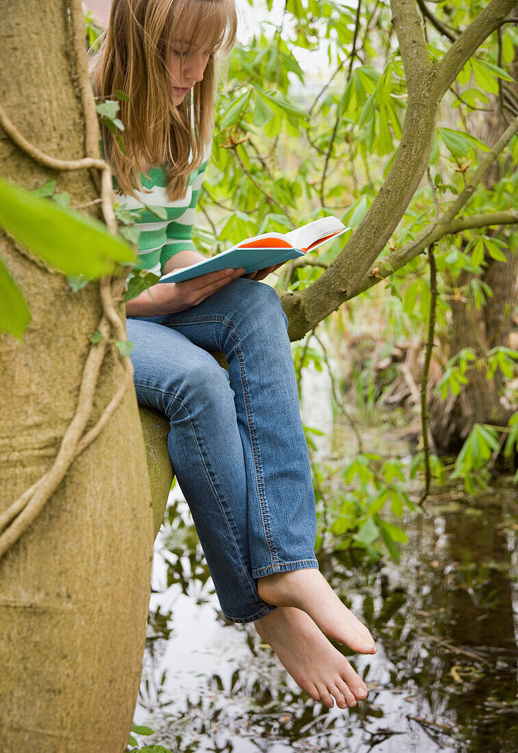 Teenage girl sitting on a the bough of a tree reading a book\n