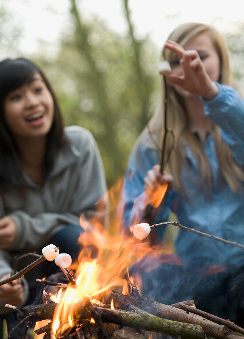 Zwei lächelnde Mädchen im Teenageralter rösten Marshmallows über einem Lagerfeuer