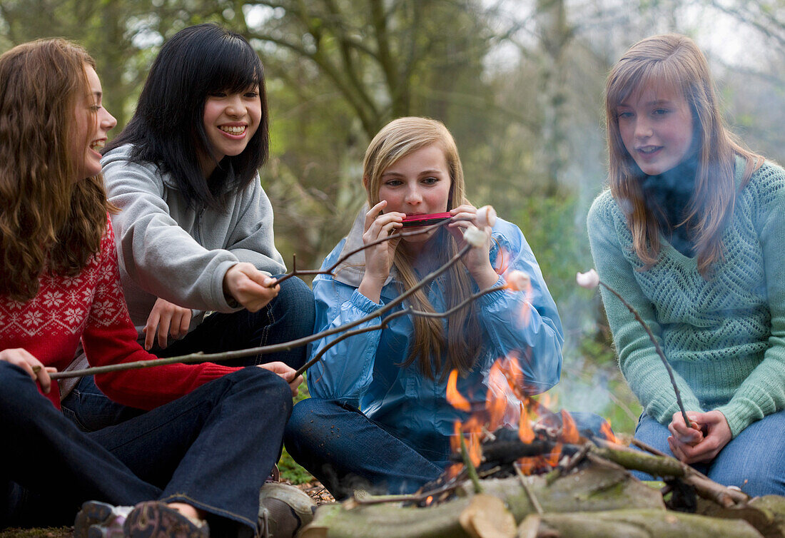 Teenage girls roasting marshmallow over campfire one is playing the harmonica\n
