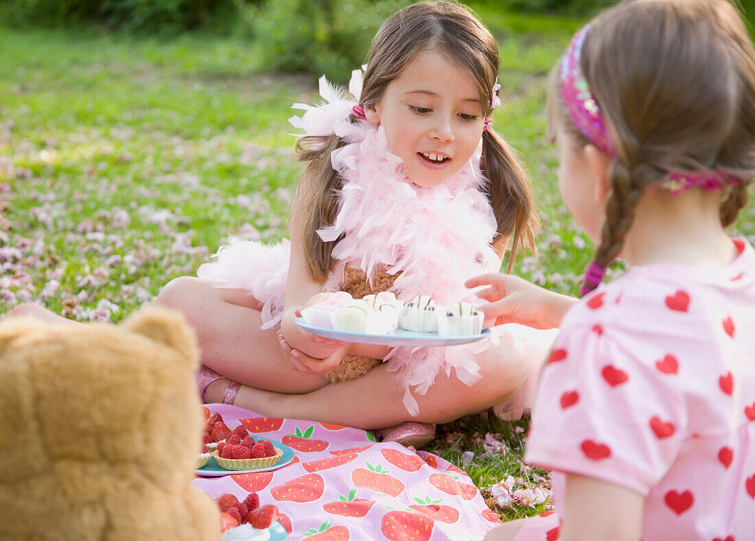 Junges Mädchen mit rosa Federboa sitzt auf einer Wiese und bietet einer Freundin Muffins an
