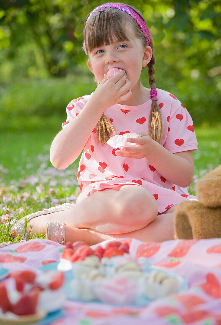 Young girl sitting in a garden smiling and eating a cupcake\n