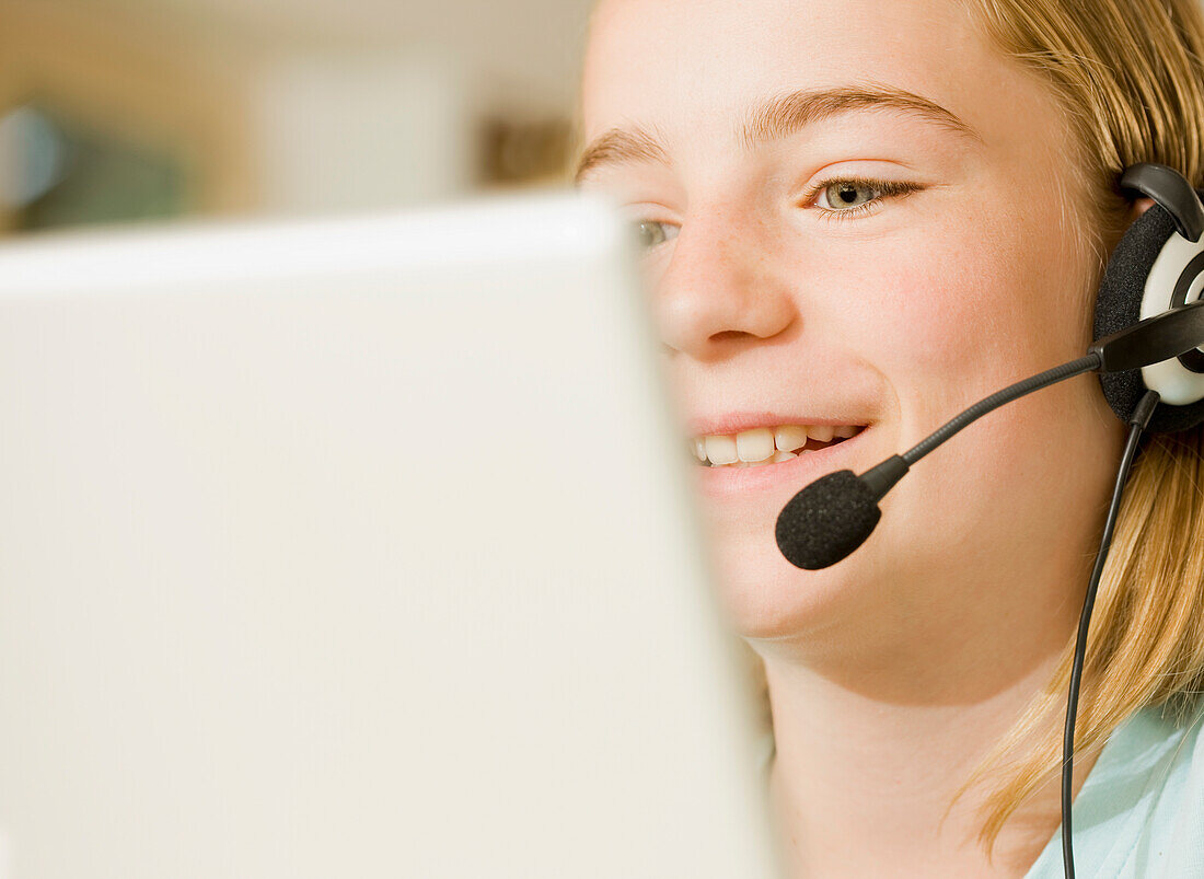 Close up of smiling young girl wearing earphones and microphone looking at laptop computer screen\n