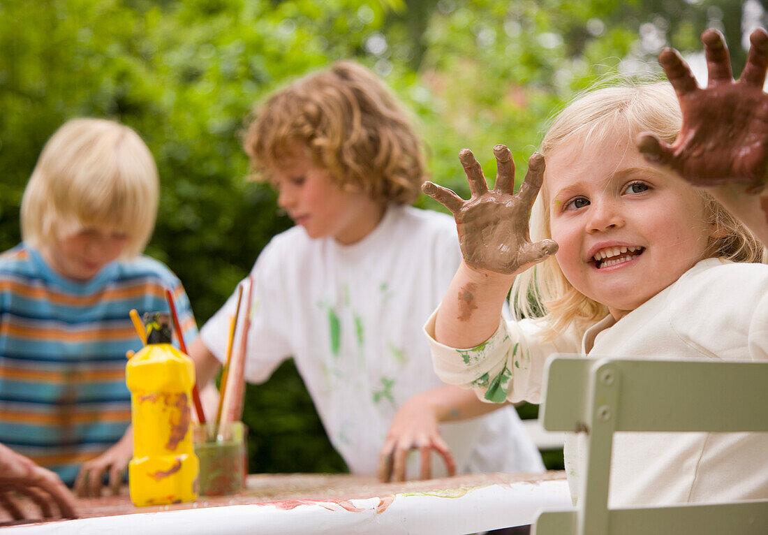Young girl sitting at table holding arms up with hands covered in paint\n