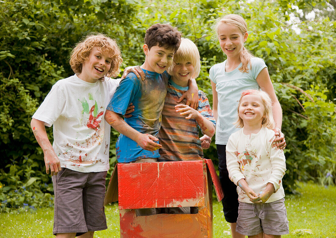 Children covered in watercolor paint in a garden, two of them standing in a cardboard box\n