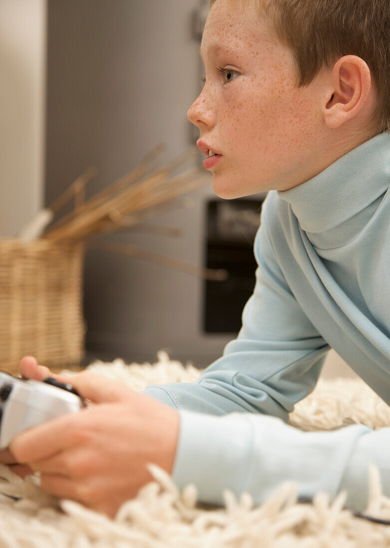 Profile of a young boy lying on a rug and holding a video game control\n