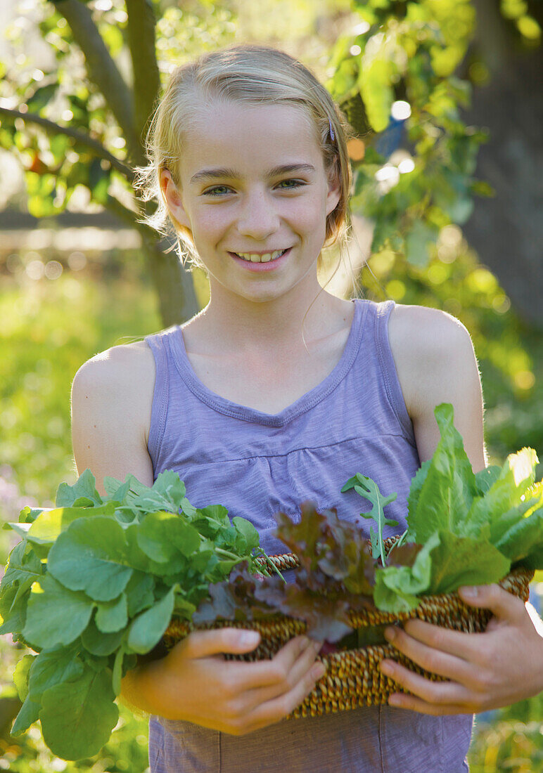 Smiling young girl standing in a garden holding a basket of leafy vegetables\n