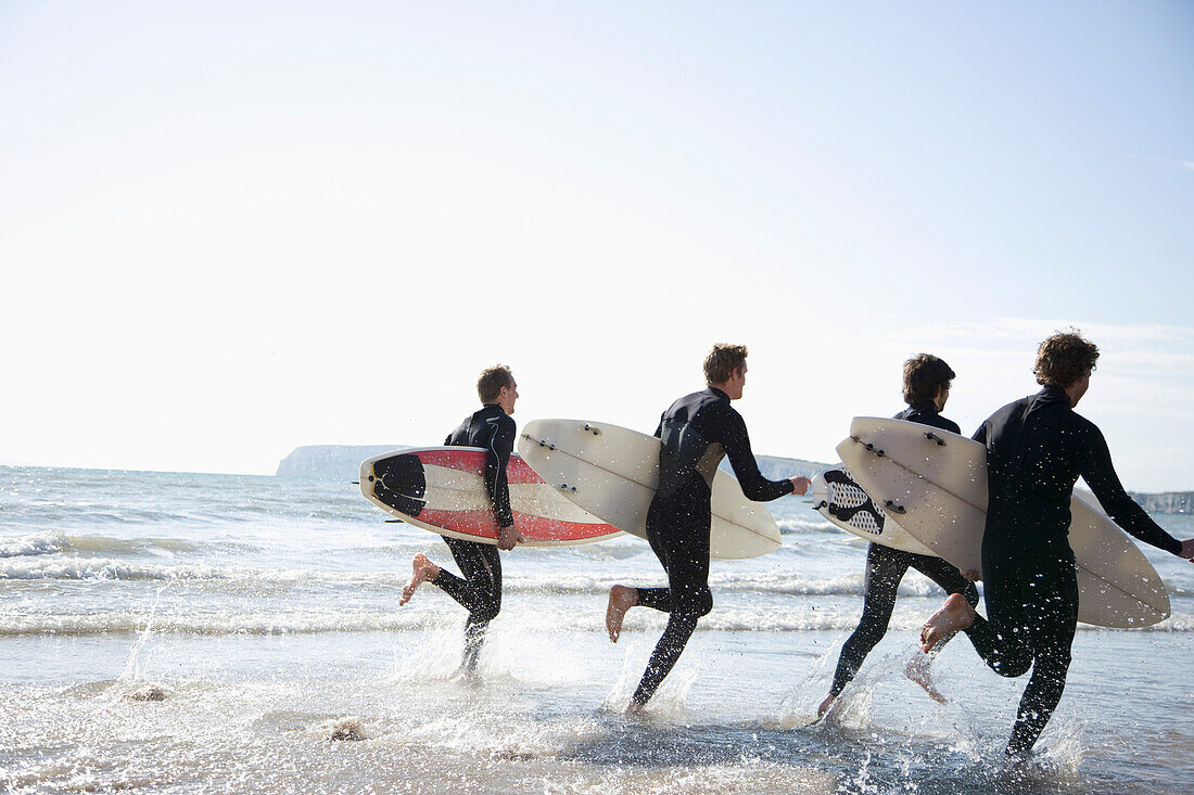 Back view of surfers running in the sea holding surfboards\n
