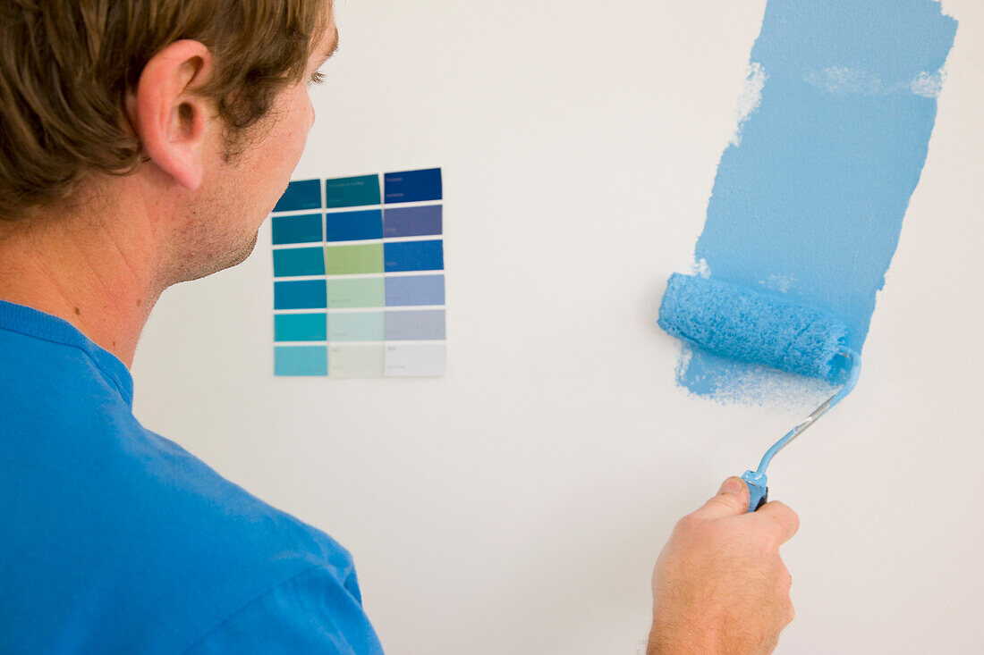 Rückenansicht eines Mannes, der einen Farbroller hält und eine weiße Wand mit blauer Farbe streicht