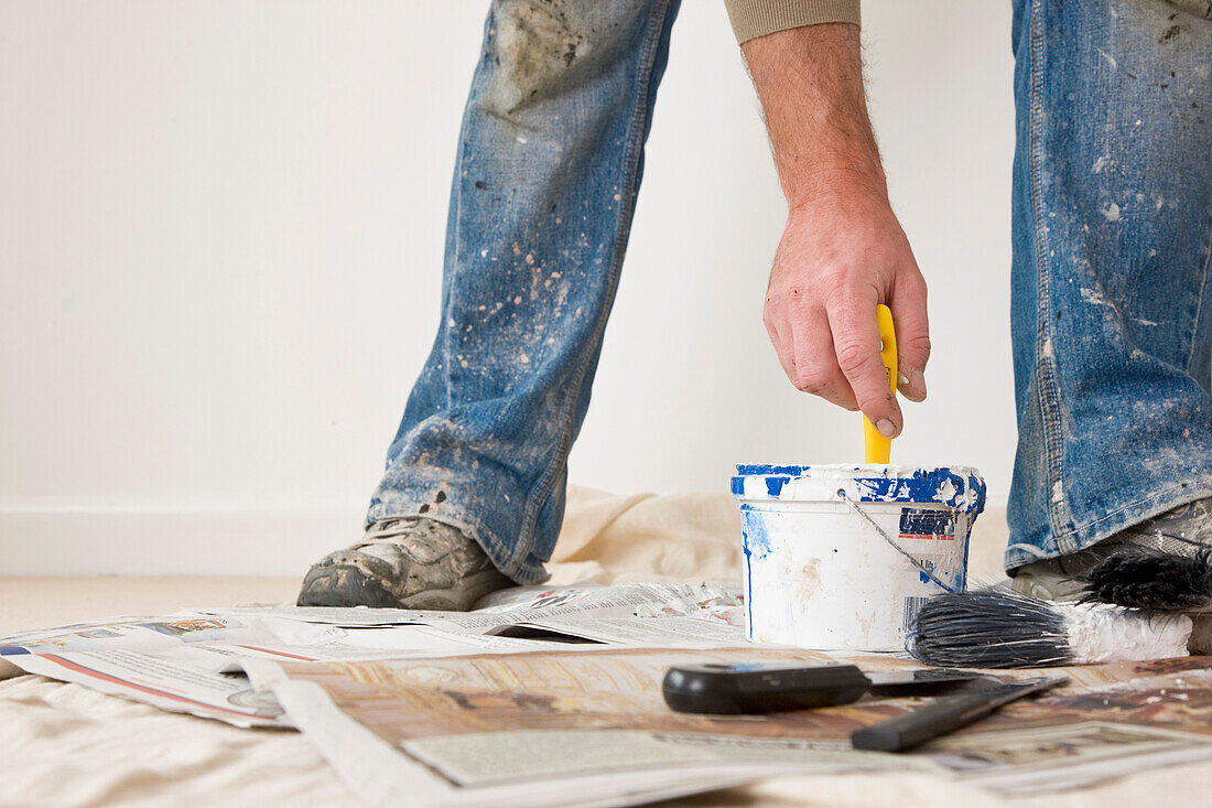 Close up of man legs wearing shoes and jeans covered in paint with his hand dipping a  paint brush\n