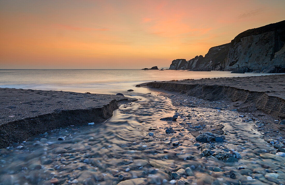 A stream cuts across sand as it reaches the sea, at dusk, in Ayrmer Cove, a remote cove near Kingsbridge, south coast of Devon, England, United Kingdom, Europe\n