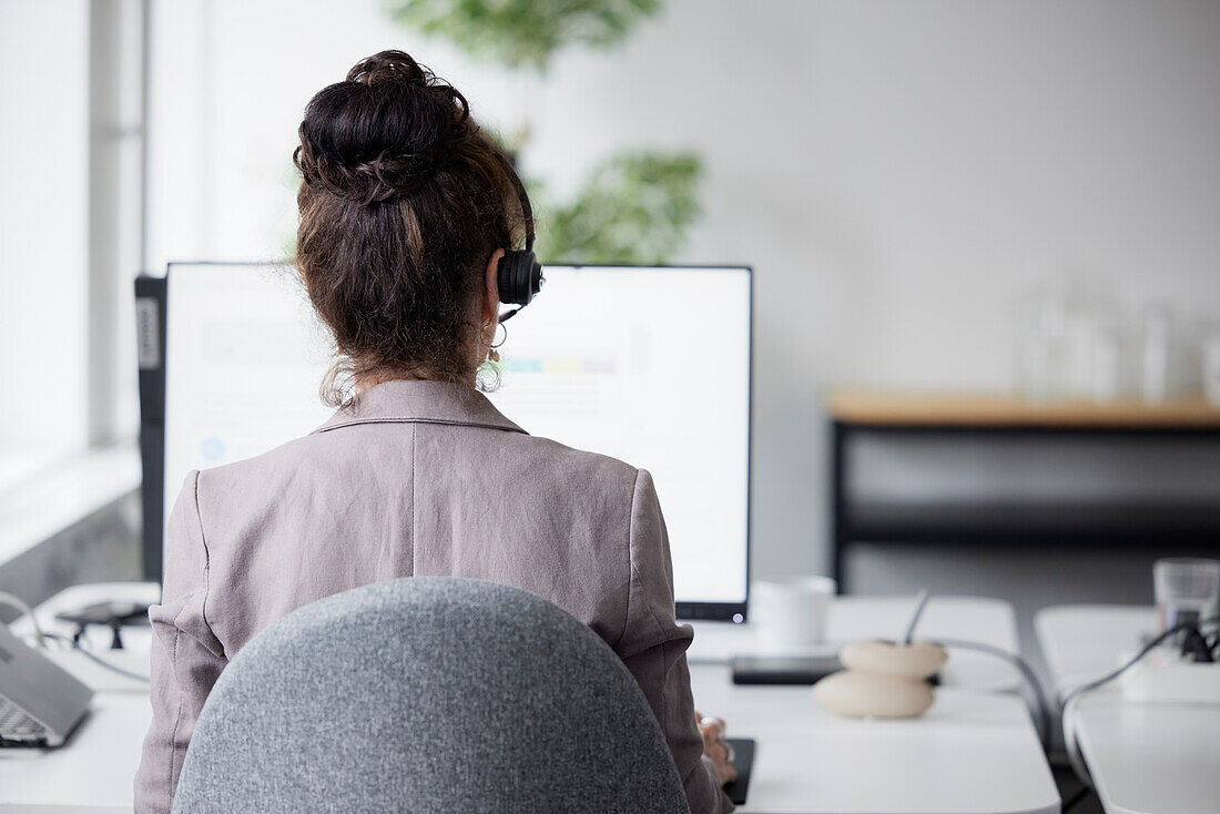 Rückansicht einer Geschäftsfrau mit Headset im Büro vor einem Computerbildschirm