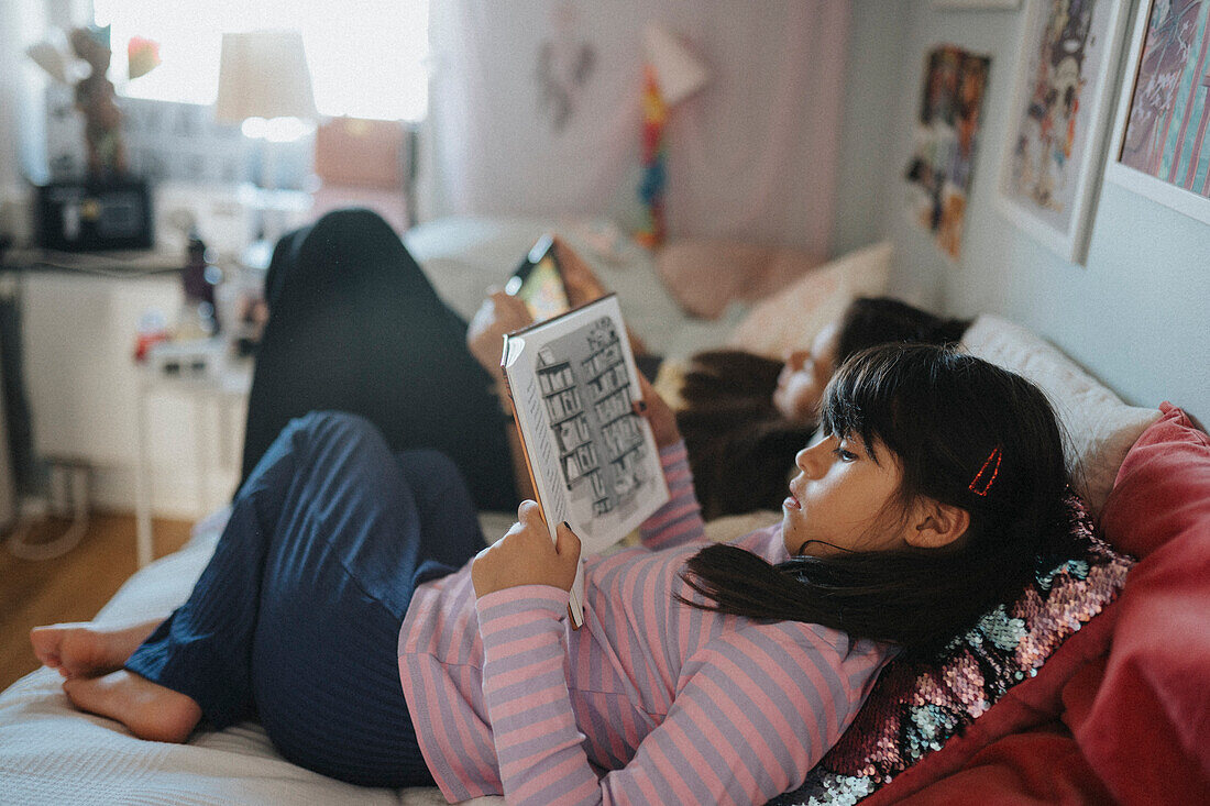 Schwestern entspannen sich im Schlafzimmer, lesen ein Buch und benutzen ein Tablet