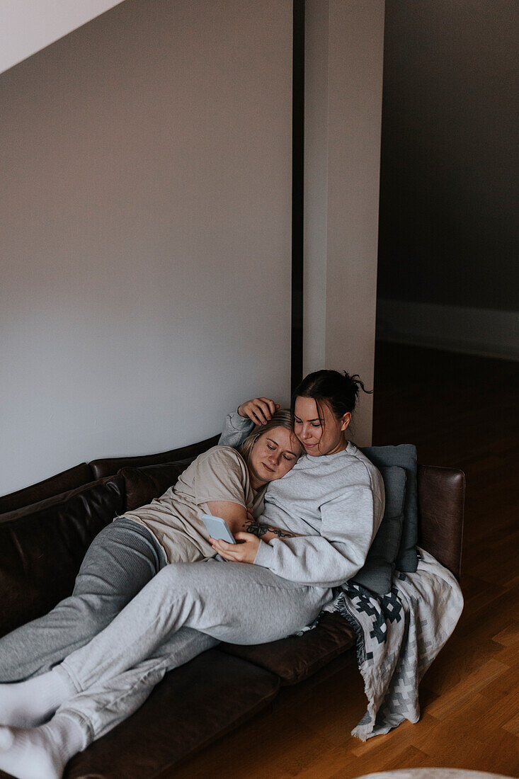 Female couple hugging together on sofa and looking at cell phone\n